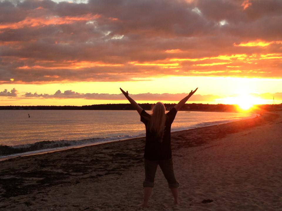 Dr. Marcia McFee standing with arms outstretched on the shoreline at sunrise